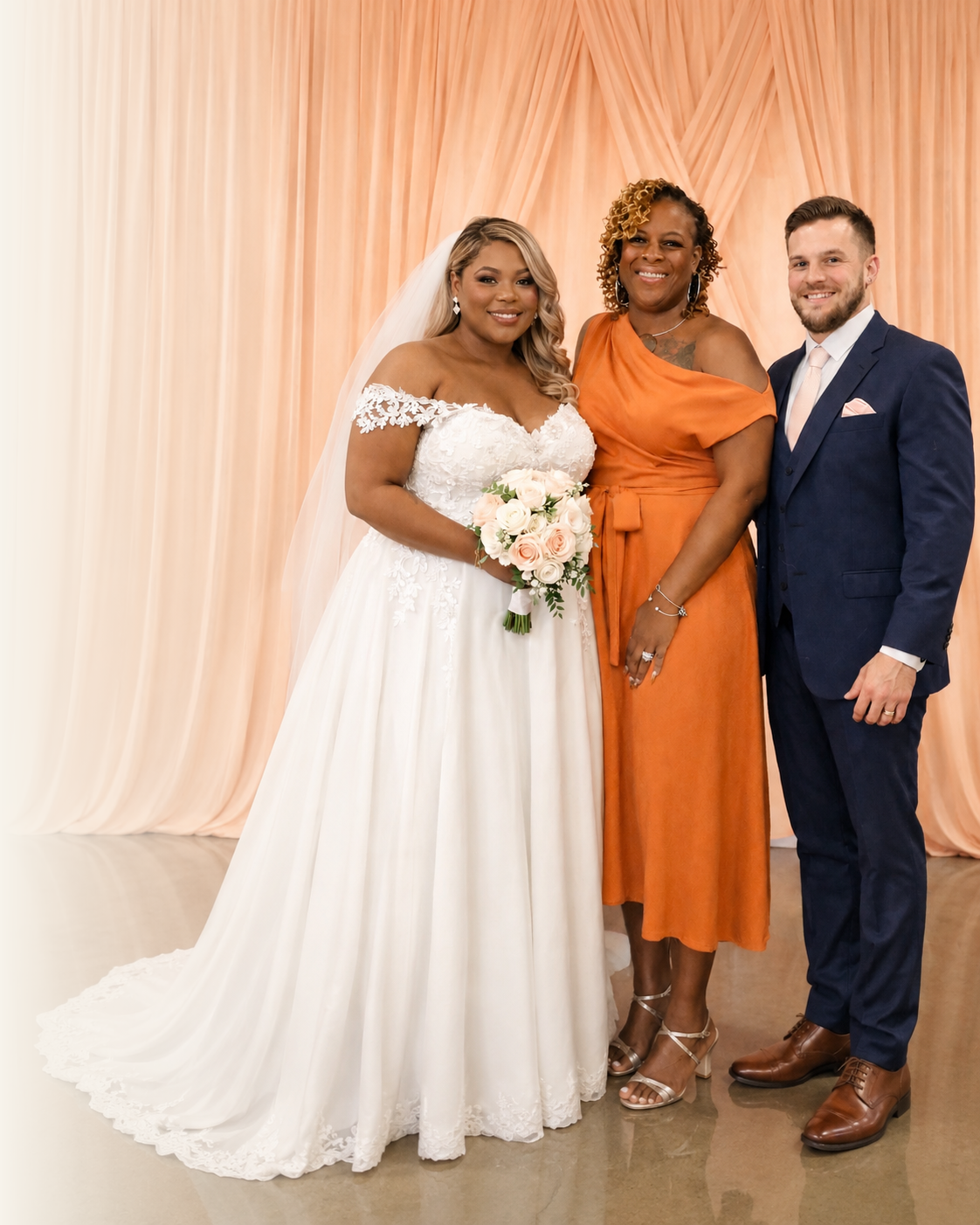 A bride and groom standing with Kesha in an orange dress against warm ivory ceremony draping